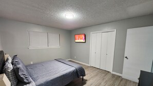 Een slaapkamer met een bed, een aan de muur bevestigde televisie, een kledingkast en een raam met jaloezieën.