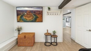 Un pasillo con un cuadro de paisaje grande, un cofre de mimbre, dos mesitas con plantas y una puerta blanca.