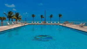 Piscina azul cristalina con sillas de descanso y palmeras, rodeada por una cerca, bajo un cielo despejado.