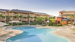 A swimming pool with a curved edge, surrounded by lounge chairs and umbrellas, with a multi-story building in the background. - 1-bedroom villa in charming Indio with AC, WiFi, fitness room (Indio)