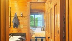 A wooden cabin interior with a bathroom featuring a sink, towel rack, and toilet paper holder. A bed with pillows and a blanket is visible through an open door.