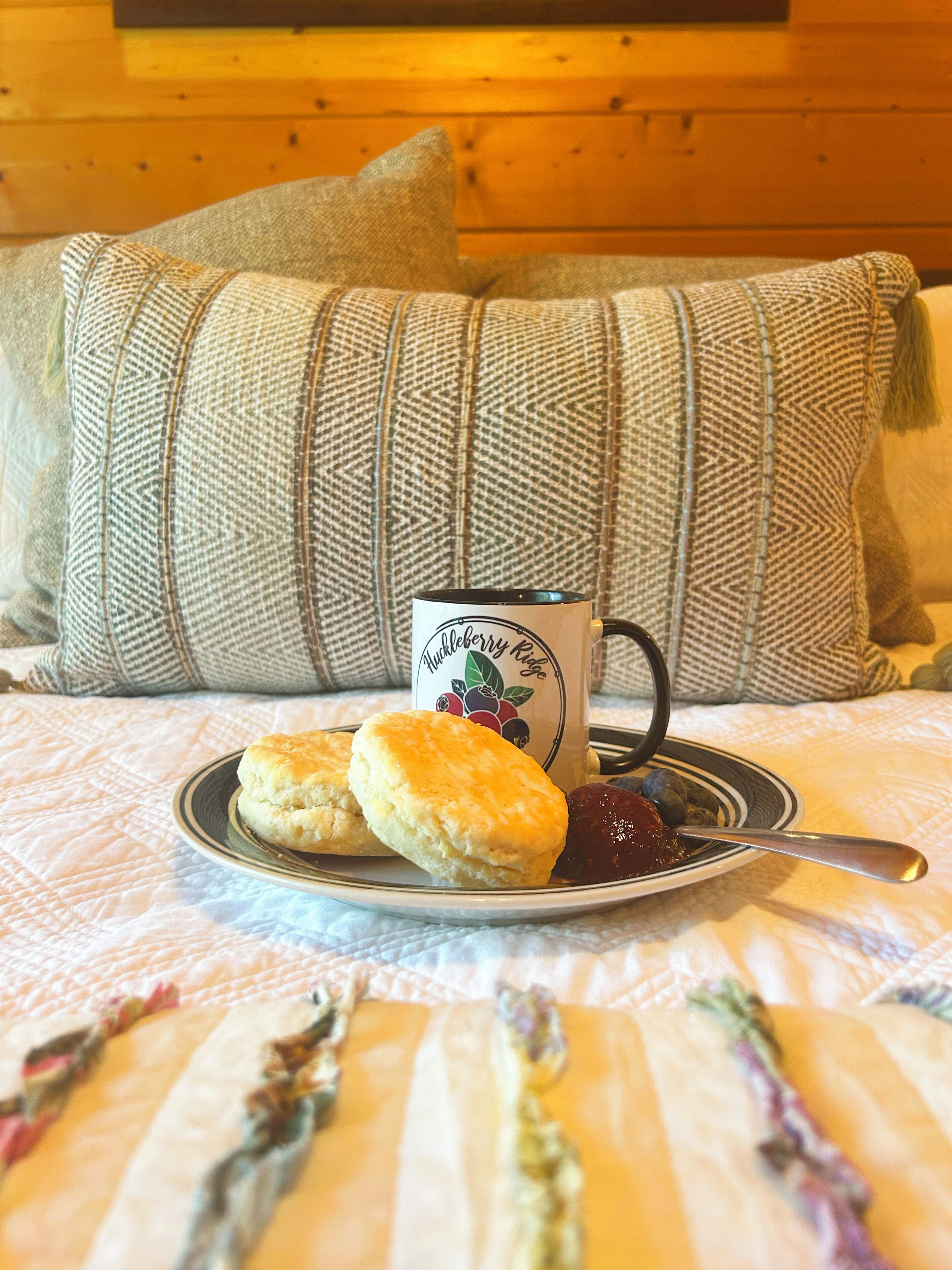 A bed with a quilted blanket, a plate with biscuits and jam, and a mug with a logo.