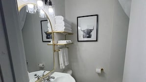 A bathroom with a white sink, toilet, and bathtub. There are towels stacked on a shelf, and a framed picture of a bull on the wall.