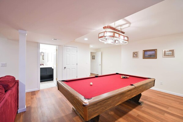A pool table with a red felt surface, a wooden frame, and a cue ball. There is a red sofa to the left and a door leading to another room.