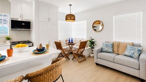 A modern kitchen with white cabinets, a central island with seating, a dining area with wooden chairs, a grey sofa, and a large window with blinds.