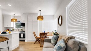 A modern kitchen with white cabinets, a dining area with a wooden table and chairs, and a cozy living area with a sofa and a large window with blinds.