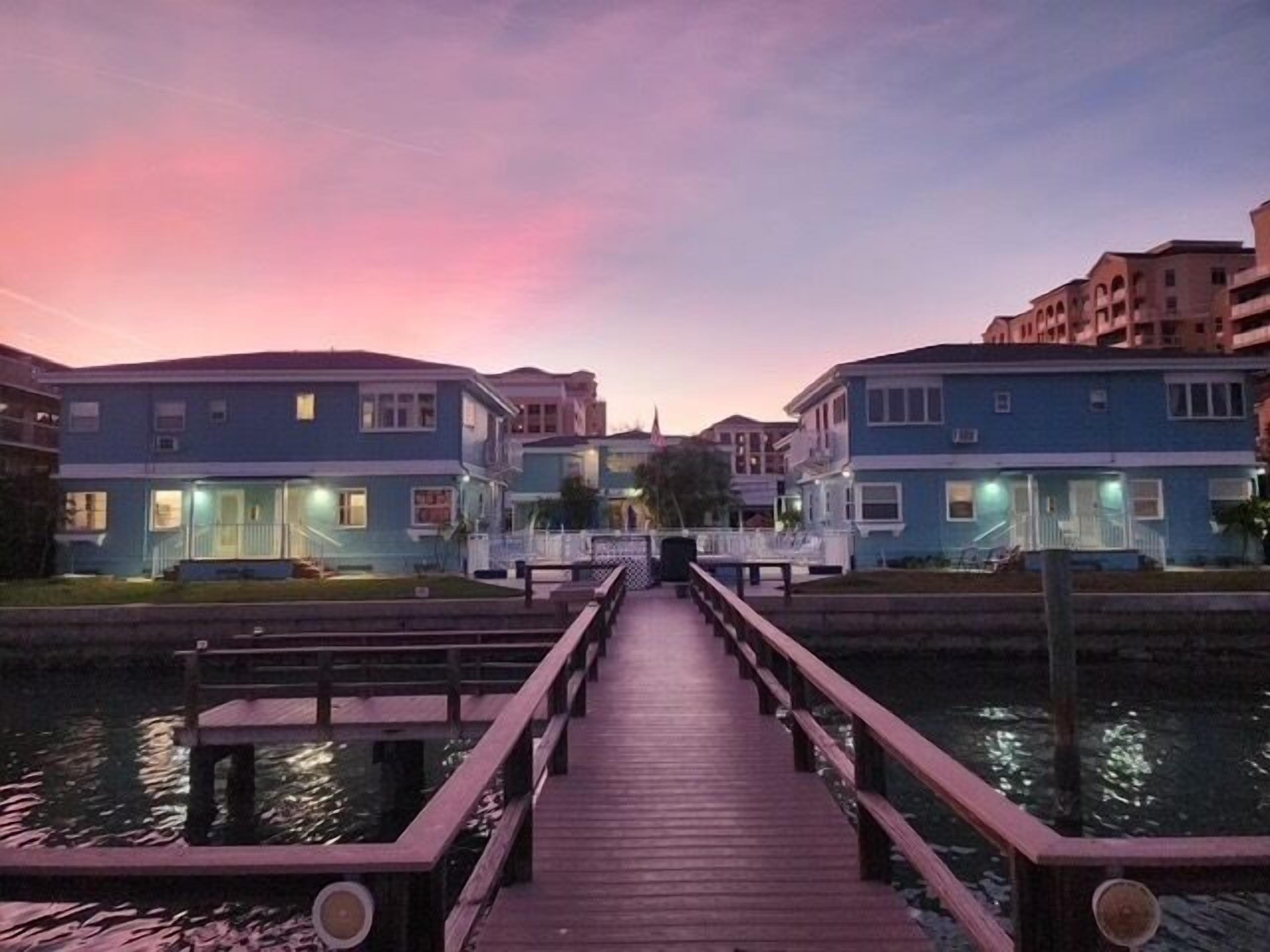 A wooden pier leading to a row of blue buildings with a sunset in the background.