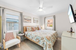 Un dormitorio con una cama, una silla, una mesita de noche, un ventilador de techo y un televisor montado en la pared.