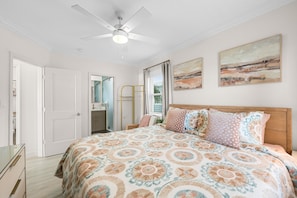 Un dormitorio con cama, ventilador de techo, ventana con cortinas y dos cuadros colgados en la pared.