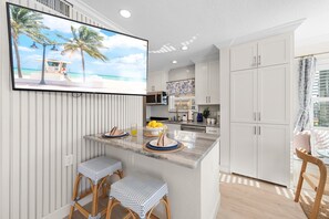 Una cocina moderna con armarios blancos, una isla central con taburetes y un televisor de pantalla plana mostrando una escena de playa.