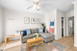 Ein modernes Wohnzimmer mit einem grauen Ecksofa, einem Holztisch, einem Deckenventilator und einem großen Fenster.