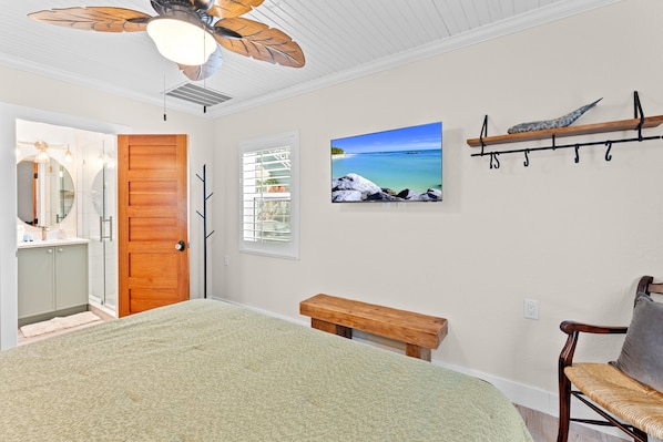 A bedroom with a bed, a wooden bench, a ceiling fan, a wall-mounted TV, and a wooden shelf with hooks.