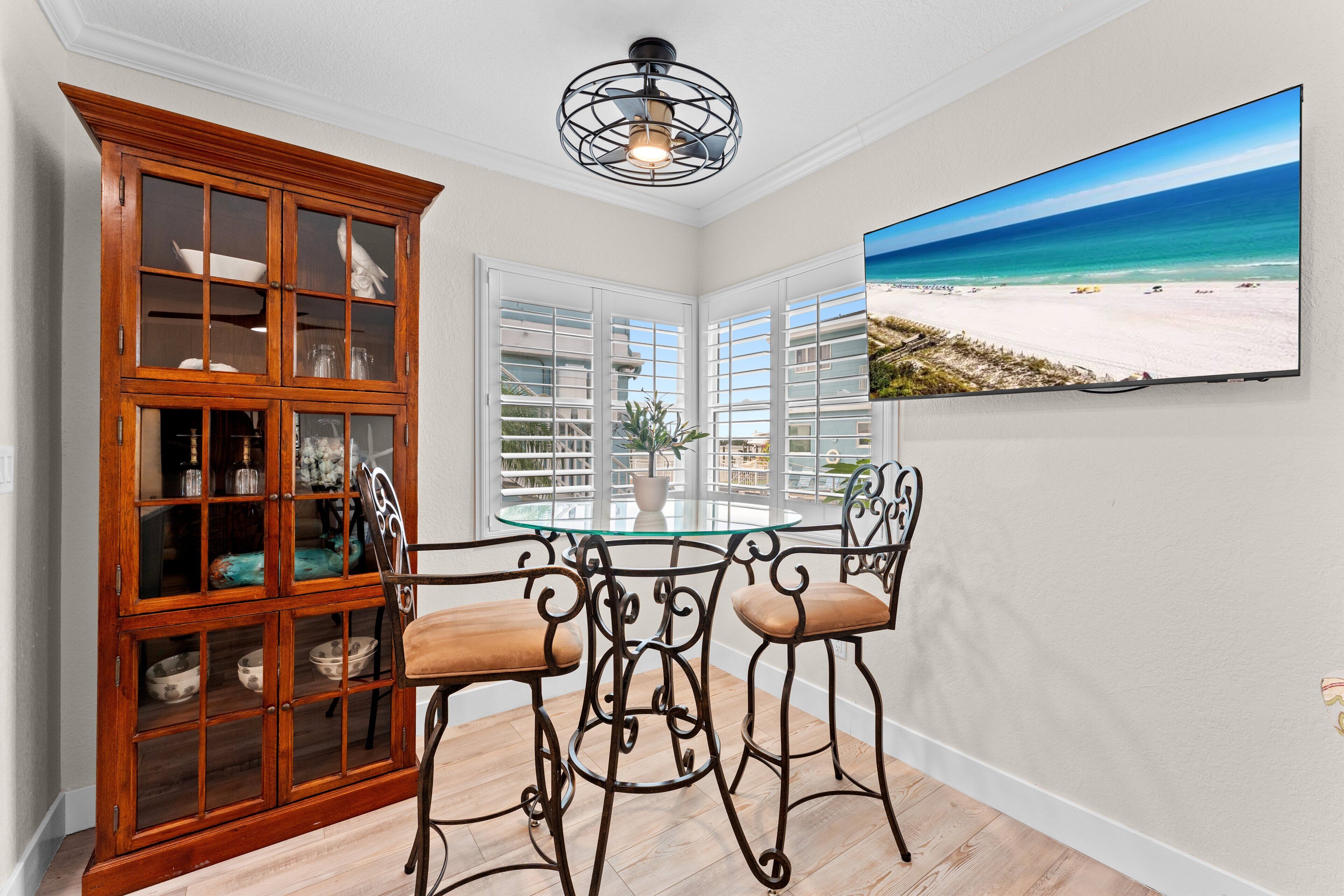 Un comedor con una mesa de vidrio, dos sillas, un armario de madera y un televisor de pared que muestra una escena de playa.