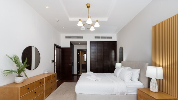 A modern bedroom with a large bed, a wooden dresser, a round mirror, and a ceiling light fixture. - 2BR at the Palm | Balcony, Pool, Gym & Beach (Dubai)