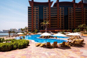 A hotel pool area with sun loungers and umbrellas, surrounded by palm trees and a view of the sea.