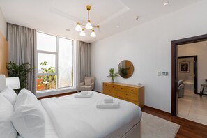 A modern hotel room with a large bed, a wooden dresser, a round mirror, and a view of the outdoors.