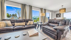 Una sala de estar moderna con sofás de cuero, una mesa de centro y grandes ventanales que ofrecen vistas a una montaña y árboles.