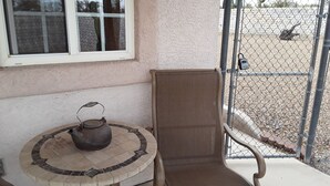 A small outdoor seating area with a metal chair and a table, a metal fence, and a window with a view of a gravel area.