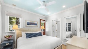 Un dormitorio con cama, lámparas de noche, ventilador de techo, ventana con persianas, puerta y televisor sobre un mueble bajo.