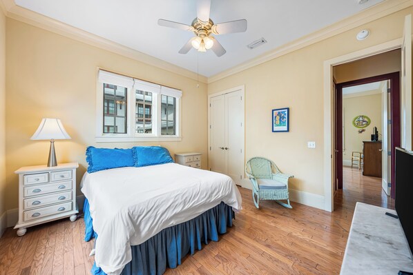 A bedroom with a bed, a nightstand, a lamp, a ceiling fan, a window, a door, a chair, and a TV.