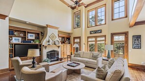 A spacious living room with a fireplace, large windows, and a high ceiling fan.