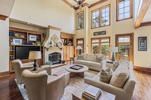 A spacious living room with a fireplace, large windows, and a high ceiling fan.