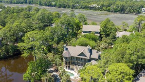 A large house with a pool, surrounded by dense trees and a body of water.