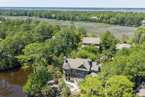 A large house with a pool, surrounded by dense trees and a body of water.