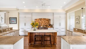 A spacious kitchen with white cabinetry, a large island with stools, a wooden backsplash, and a ceiling fan.