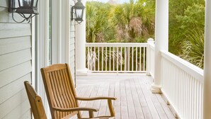 A wooden porch with two chairs and two wall-mounted lanterns, overlooking a lush green landscape.