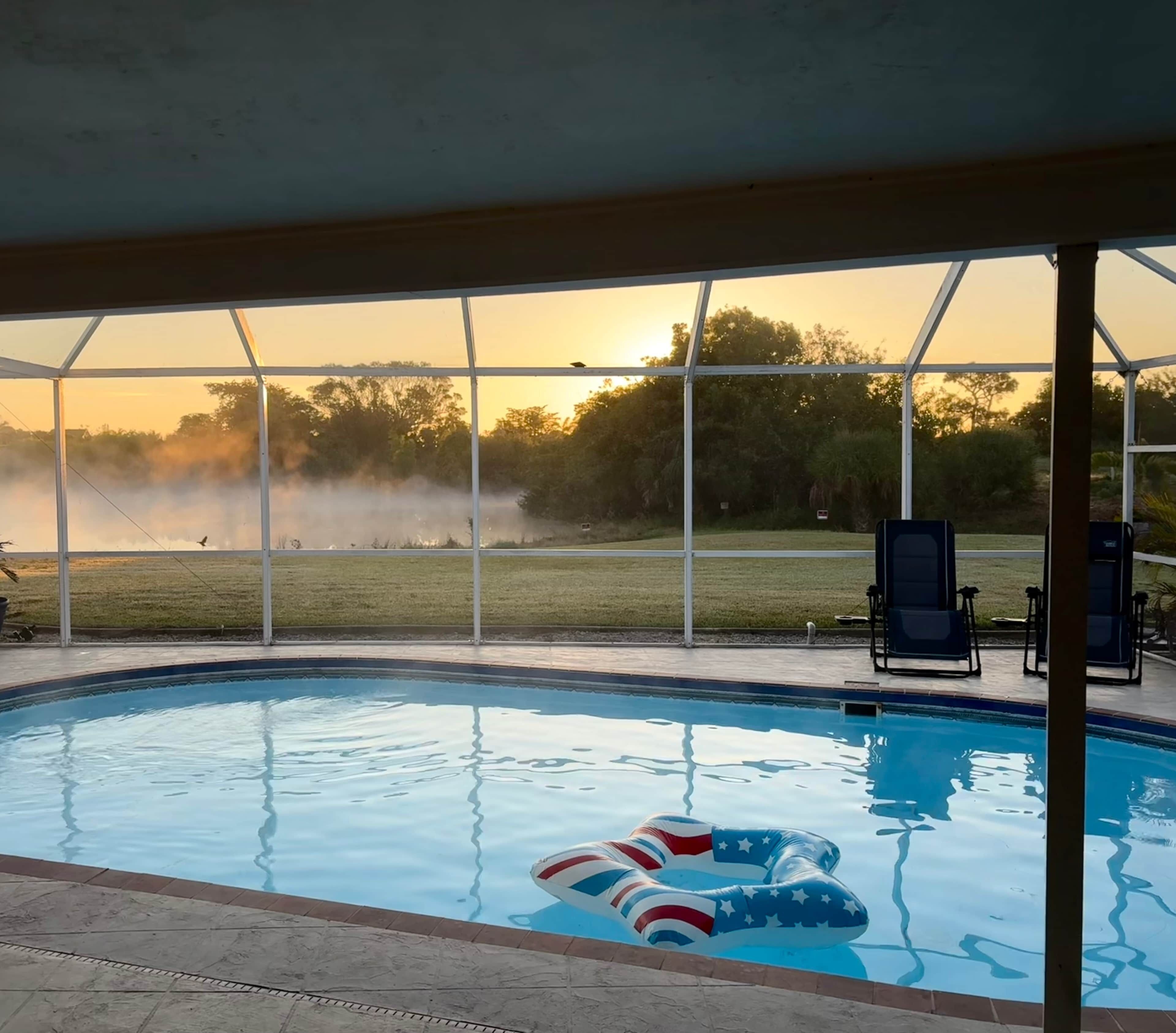 A pool with a view of a field and trees, surrounded by a covered area with chairs.