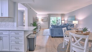A modern living room with a blue sofa, a dining table with chairs, and a kitchen area with white cabinets and a trash bin. - Renovated 1BR Condo. Walk to Beach&Salty Dog, Sleeps 5. Bike, Golf, Pickleball! (Hilton Head Island)
