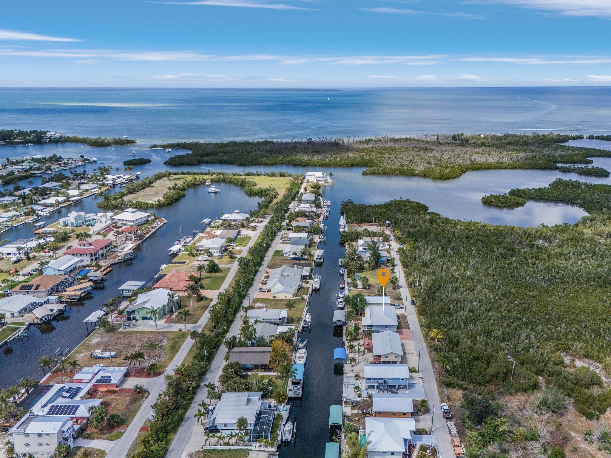 An aerial view of a coastal residential area with houses along a narrow waterway, surrounded by mangroves and open water.