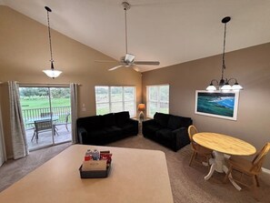 A room with a ceiling fan, a table with chairs, a black sofa, a window with blinds, and a view of a green landscape.