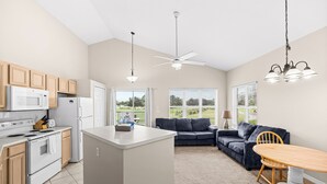 A modern kitchen with a central island, a dining area with a wooden table, a dark blue sofa, and a ceiling fan. - Right Direction Rentals 1 Bedroom Villa 9906 (Port St. Lucie)