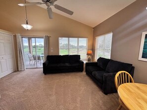 A living room with a ceiling fan, two black sofas, a wooden table, and a view of a green landscape through the windows.