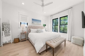 Une chambre à coucher avec un grand lit, une table de chevet, une commode, un banc, un ventilateur de plafond et une fenêtre avec des rideaux.