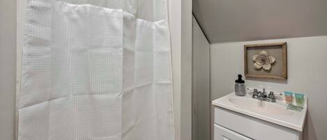 Un baño con cortina de ducha, un lavamanos con artículos de aseo y un cuadro en la pared.