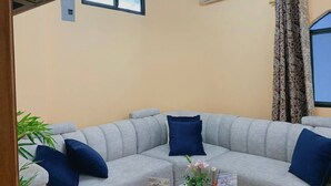 Ein Wohnzimmer mit grauem Sofa, blauen Kissen, einem kleinen Tisch mit einer Vase und einem Fenster mit Vorhängen.