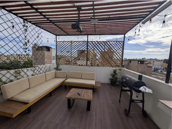 A rooftop patio with a wooden bench, a sofa, a small table, and a grill.