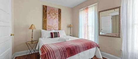 Un dormitorio con cama, dos mesitas de noche, ventilador de techo, espejo grande y ventana con cortinas.