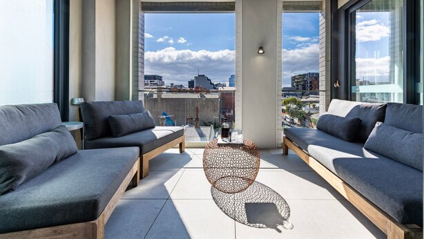 A modern balcony with grey sofas and a wooden bench, a woven coffee table, and a city view.