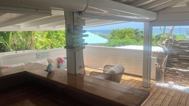 Una terraza de madera con un banco y sillas, con vistas a un jardín exuberante y el océano.