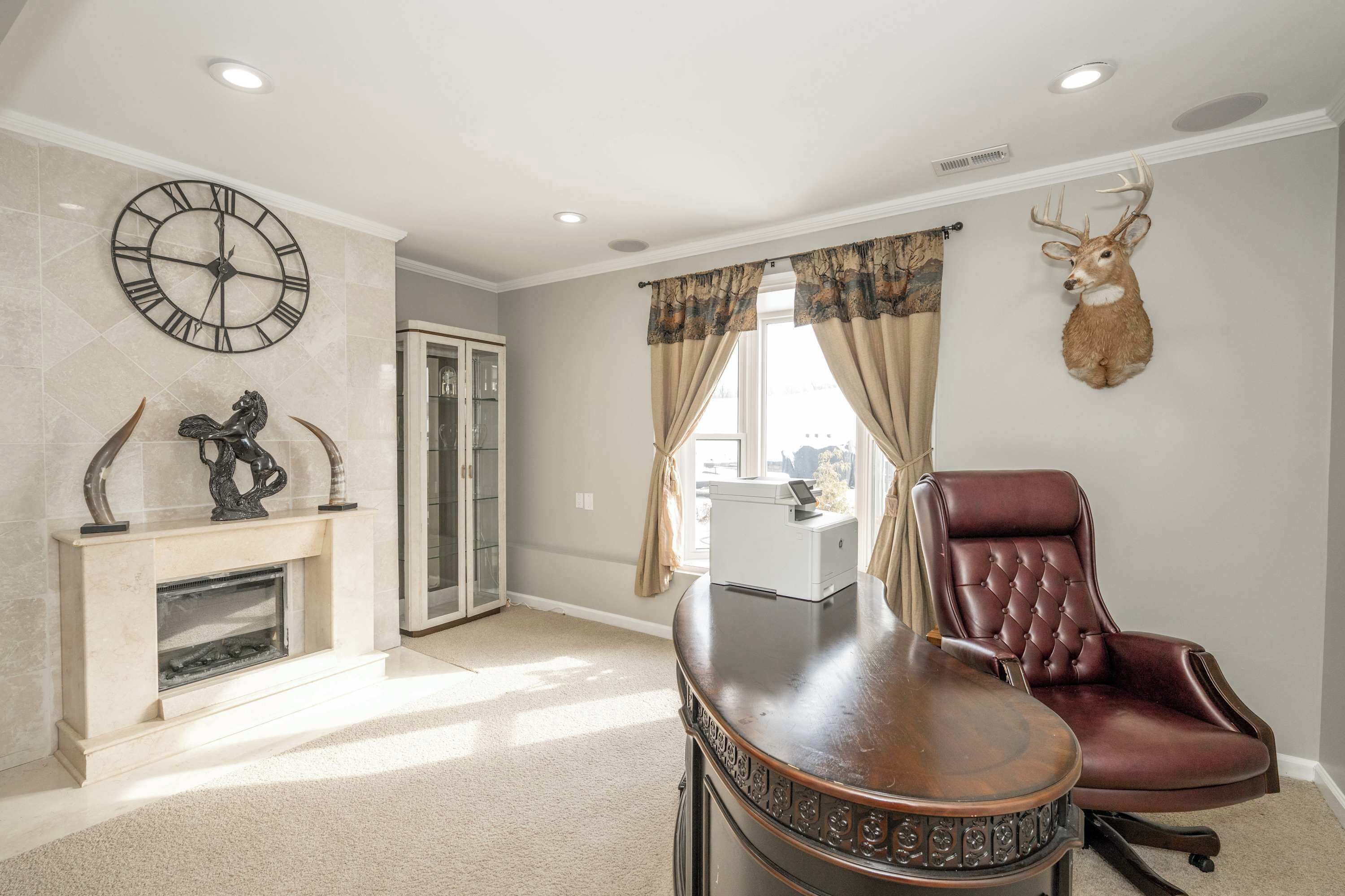 A spacious room with a fireplace, a large clock, a deer head mounted on the wall, and a leather chair.