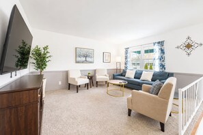Ein Wohnzimmer mit Flachbildfernseher, einer Couch, einem Sessel, einem Couchtisch und einer Pflanze.