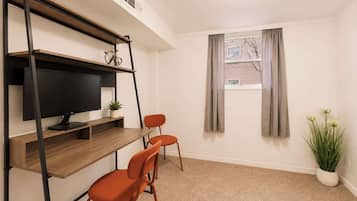 Una habitación con escritorio, dos sillas, televisor, ventana con cortinas y planta en maceta.