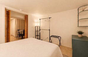 A hotel room with a bed, a desk, a chair, a wardrobe, a hanging rack, and a small plant. - The Gahanna Getaway (Columbus)