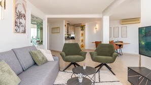 Un salon moderne avec un canapé gris, deux fauteuils verts, une petite table basse et un téléviseur posé sur un meuble.