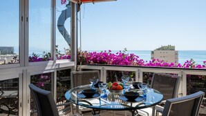 Une table en verre, des chaises et un saladier de fruits sur un balcon offrant une vue sur la mer et les bâtiments.