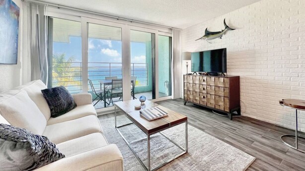 Un salon moderne avec un canapé, une table basse et un meuble pour la télévision. Il y a un balcon avec vue sur l’océan.
